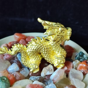 May include: A detailed, golden dragon figurine with intricate scales and a dynamic pose. The dragon is set on a bed of colorful, polished stones, creating a striking contrast. The figurine's metallic sheen catches the light, highlighting its features.
