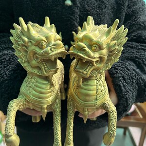 May include: Two golden-colored, mythical creature statues. Each statue has a dragon-like head with open mouths, spiky manes, and detailed scales. The statues are held up by a person.
