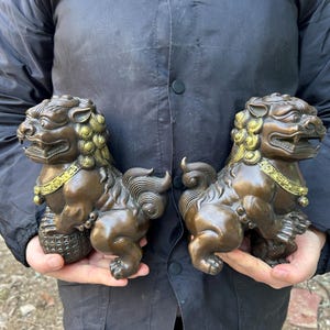 May include: Two bronze Foo Dog statues, each held in a person's hands. The statues have detailed features, including curled manes, open mouths, and decorative collars. They are sitting on their haunches, with one paw resting on a ball.