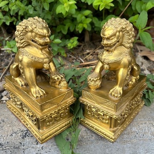 May include: Two golden lion statues, each seated atop a rectangular base with intricate carvings. The lions have detailed manes and fierce expressions. The statues are made of a shiny, metallic material, and are displayed outdoors.