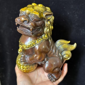 May include: A bronze-toned Foo Dog statue featuring a polished gold-colored mane, tail, and collar. The mythical creature is depicted with its mouth open, displaying teeth, and one front paw resting on a textured, spherical gold-colored ball.