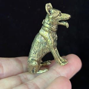 Puede incluir: Pequeña y detallada figura de latón de un perro sentado con la lengua fuera. El perro tiene un collar y está hecho de metal dorado. La figura se sostiene en la mano de una persona sobre un fondo negro.
