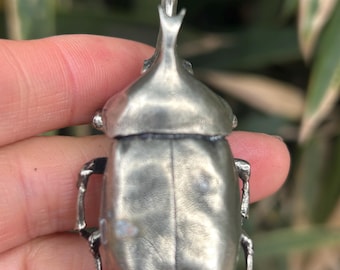 Könnte beinhalten: Eine silberfarbene Käferfigur aus Metall mit einem markanten Y-förmigen Horn am Kopf. Das Insektenmodell hat eine glatte, reflektierende Oberfläche, einen segmentierten Körper und dünne, gegliederte Beine an den Seiten des Brustkorbs.