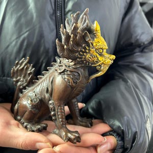 May include: A bronze-colored, mythical creature figurine with gold accents. The detailed sculpture features a dragon-like body, a lion's mane, and a fierce expression. The figurine is held in two hands, showcasing its intricate design and craftsmanship.