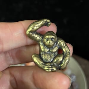 May include: A small, bronze-colored figurine of a seated monkey with one arm raised. The monkey has detailed facial features and is sitting with its legs crossed. The figurine is held in a person's hand.