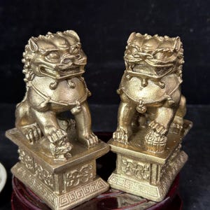 May include: A pair of gold-colored brass guardian lion statues, known as Shishi, each mounted on a rectangular pedestal with intricate geometric carvings. One lion rests a paw on a small cub, while the other rests a paw on a decorative ball.