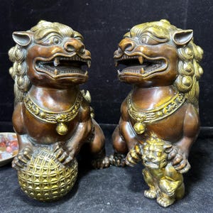 May include: Two bronze-colored guardian lion statues with gold accents. One lion holds a patterned ball, the other has a smaller lion cub. Both lions have detailed manes, collars, and open mouths, showcasing their teeth.