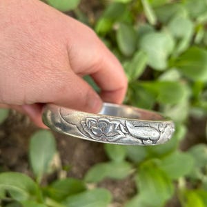 May include: A silver-toned bangle bracelet with floral and fish motifs. The bracelet has a detailed, embossed design featuring a lotus flower and a fish. The bracelet is held against a backdrop of green foliage.