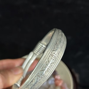 May include: A silver bangle bracelet featuring detailed floral and leaf patterns. The bracelet has a textured surface and a classic design. The bangle is a piece of jewellery.