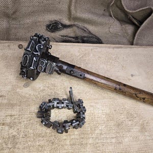Op de afbeelding: Een vintage metalen stempel met de nummers "0180" en de letters "TS" op de stempelkop, bevestigd aan een houten handvat. Er is ook een cirkelvormige metalen stempel aanwezig. De items liggen op een lichtbruin houten oppervlak.