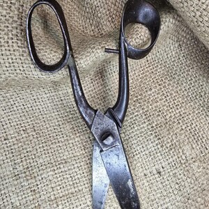 May include: Antique metal scissors with large finger loops and pointed blades. The scissors have a dark, aged appearance, showing signs of wear. The blades are slightly open, suggesting they are ready for use. The scissors are set against a textured, tan background.