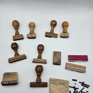 Könnte beinhalten: Eine Sammlung von Vintage-Holzstempeln mit abgerundeten Griffen. Die Stempel haben rechteckige Basen mit verschiedenen Texteindrücken. Ebenfalls enthalten sind Stempelkissen und ein kleines Papierpäckchen mit handschriftlichem Text und kleinen schwarzen Tintenstreifen.