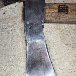 May include: Close-up of a vintage axe head with a wooden handle. The axe head is made of dark metal and shows signs of wear. The wooden handle has a stamp. The axe is positioned on a light-colored wooden surface.