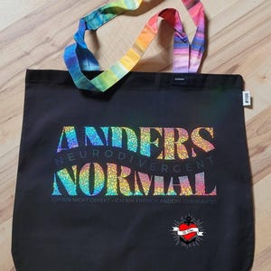 May include: Black tote bag with the words "ANDERS NEURODIVERGENT NORMAL" in a colourful, glittery font. The bag has rainbow-coloured handles and the text "ICH BIN NICHT DEFEKT - ICH BIN EINFACH ANDERS VERDRAHTET".