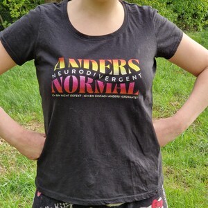 May include: Black crew neck t-shirt featuring the words "ANDERS NEURODIVERGENT NORMAL" in a gradient of yellow, orange, and pink. The phrase "ICH BIN NICHT DEFEKT - ICH BIN EINFACH ANDERS VERDRAHTET" is printed below the main text in a smaller font.
