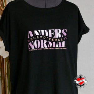 May include: Black t-shirt featuring the text "ANDERS NEURODIVERGENT NORMAL" in a pink gradient. The phrase "ICH BIN NICHT DEFEKT - ICH BIN EINFACH ANDERS VERDRAHTE" is below. A small logo is visible in the bottom right corner of the shirt.