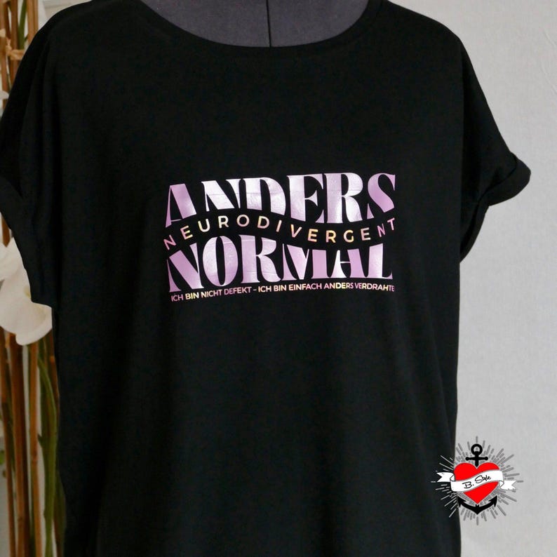May include: Black t-shirt featuring the text "ANDERS NEURODIVERGENT NORMAL" in a pink gradient. The phrase "ICH BIN NICHT DEFEKT - ICH BIN EINFACH ANDERS VERDRAHTE" is below. A small logo is visible in the bottom right corner of the shirt.