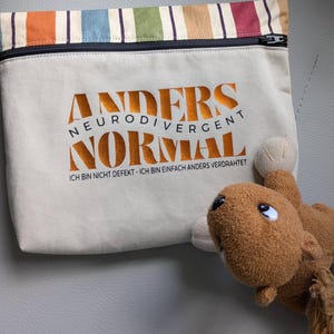 May include: A beige pouch with a black zip and the words "ANDERS NEURODIVERGENT NORMAL" in orange. The pouch has a striped top with orange, green, blue, and red stripes. A brown stuffed toy is in the foreground.