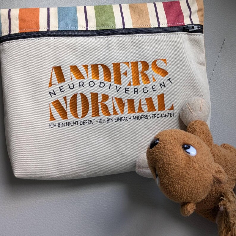 May include: A beige pouch with a black zip and the words "ANDERS NEURODIVERGENT NORMAL" in orange. The pouch has a striped top with orange, green, blue, and red stripes. A brown stuffed toy is in the foreground.