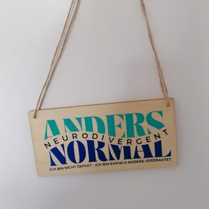 May include: A rectangular wooden sign with the words "ANDERS NEURODIVERGENT NORMAL" in teal and blue. The sign also includes the text "ICH BIN NICHT DEFEKT - ICH BIN EINFACH ANDERS VERDRAHTET" in black. The sign is suspended by two brown strings.