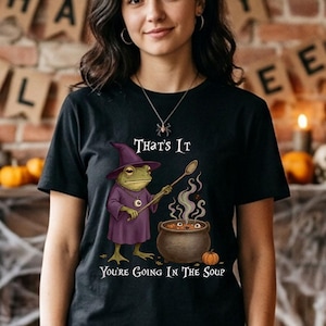 May include: Black t-shirt with a Halloween-themed graphic. The design features a frog dressed as a witch stirring a cauldron, with the text "That's It, You're Going In The Soup". The shirt is worn by a person.