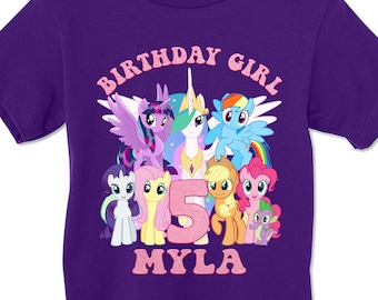 May include: A purple short-sleeve t-shirt featuring a graphic of My Little Pony characters, a large pink glittery number 5, and the text "BIRTHDAY GIRL" and "MYLA" in pink lettering. The design includes Twilight Sparkle, Celestia, Rainbow Dash, Rarity, Fluttershy, Applejack, Pinkie Pie, and Spike.