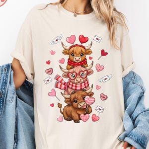May include: Ivory-colored t-shirt featuring a stacked design of three cartoon Highland cows, each adorned with Valentine's Day accessories like scarves, glasses, and heart-shaped lollipops. The design includes hearts and envelopes. The word "Ivory" is in the top left corner.