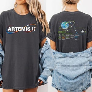 May include: Dark gray t-shirt with the text "ARTEMIS I" in a stylized design on the front. The back of the shirt features a graphic of Earth, the moon, and a flight path diagram, with the text "ARTEMIS II".