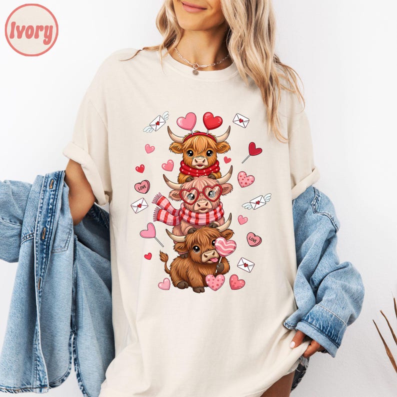 May include: Ivory-colored t-shirt featuring a stacked design of three cartoon Highland cows, each adorned with Valentine's Day accessories like scarves, glasses, and heart-shaped lollipops. The design includes hearts and envelopes. The word "Ivory" is in the top left corner.