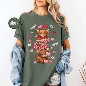 May include: Moss green t-shirt featuring a stack of three cartoon Highland cows. The cows are wearing scarves, glasses, and holding heart-shaped lollipops. Pink hearts and envelopes with hearts are scattered around the cows.