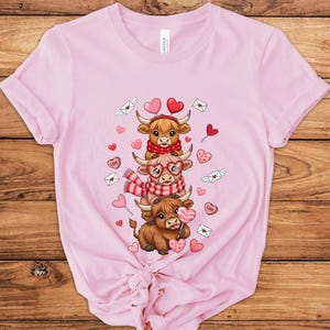 May include: Pink t-shirt featuring a graphic of three cartoon Highland cows stacked on top of each other. The cows are decorated with hearts, scarves, and glasses. The design includes hearts, love letters, and candy hearts.