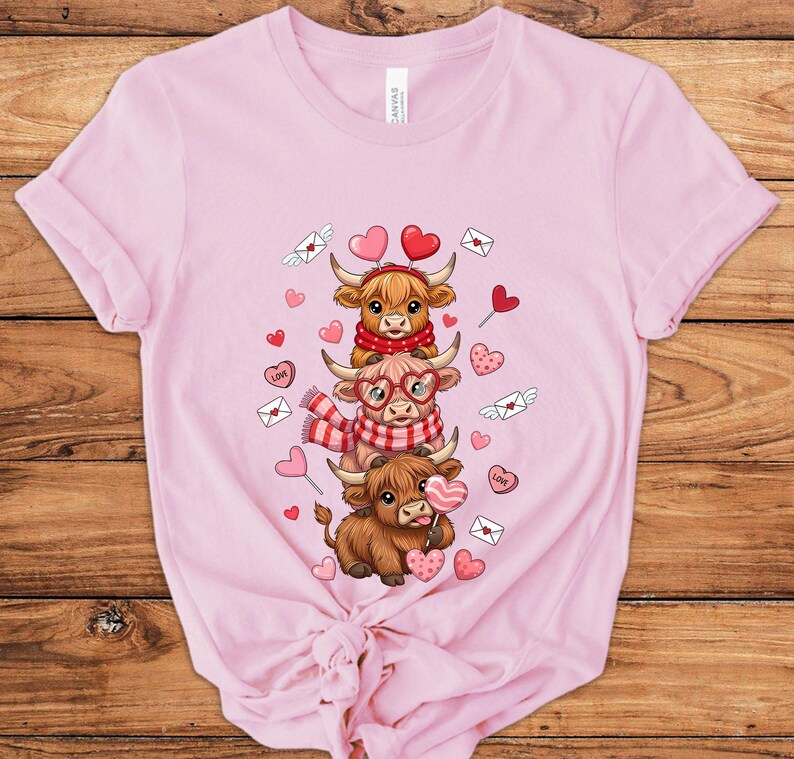 May include: Pink t-shirt featuring a graphic of three cartoon Highland cows stacked on top of each other. The cows are decorated with hearts, scarves, and glasses. The design includes hearts, love letters, and candy hearts.