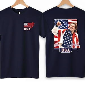 K&ouml;nnte beinhalten: Marineblaue T-Shirts mit patriotischem Thema. Eines zeigt eine stilisierte amerikanische Flagge und das Wort "USA". Das andere zeigt ein gerahmtes Bild einer Person, die in eine amerikanische Flagge geh&uuml;llt ist, mit dem Wort "USA" unten.