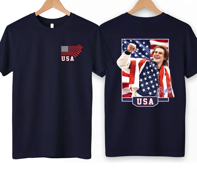 K&ouml;nnte beinhalten: Marineblaue T-Shirts mit patriotischem Thema. Eines zeigt eine stilisierte amerikanische Flagge und das Wort "USA". Das andere zeigt ein gerahmtes Bild einer Person, die in eine amerikanische Flagge geh&uuml;llt ist, mit dem Wort "USA" unten.