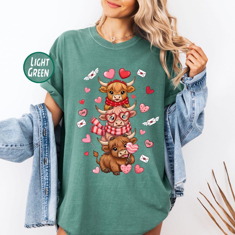 May include: Light green t-shirt featuring a stacked design of three Highland cows with heart-shaped accents. The cows wear scarves, glasses, and headbands. The shirt has the text "LIGHT GREEN" in a circular badge.