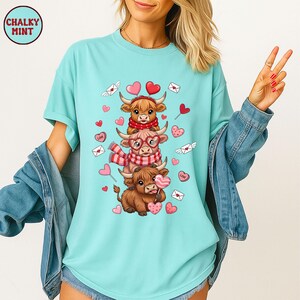 May include: A light blue t-shirt featuring a stacked design of three Highland cows, each adorned with scarves, glasses, and heart-shaped lollipops. The design includes hearts, envelopes, and the text "LOVE". The shirt has short sleeves and a relaxed fit.