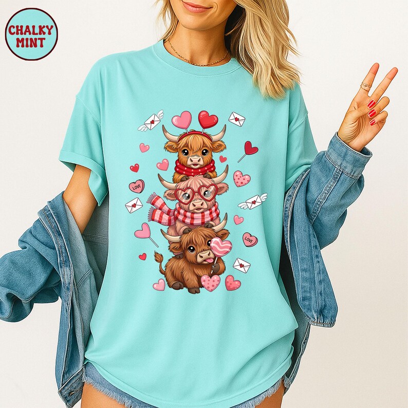 May include: A light blue t-shirt featuring a stacked design of three Highland cows, each adorned with scarves, glasses, and heart-shaped lollipops. The design includes hearts, envelopes, and the text "LOVE". The shirt has short sleeves and a relaxed fit.