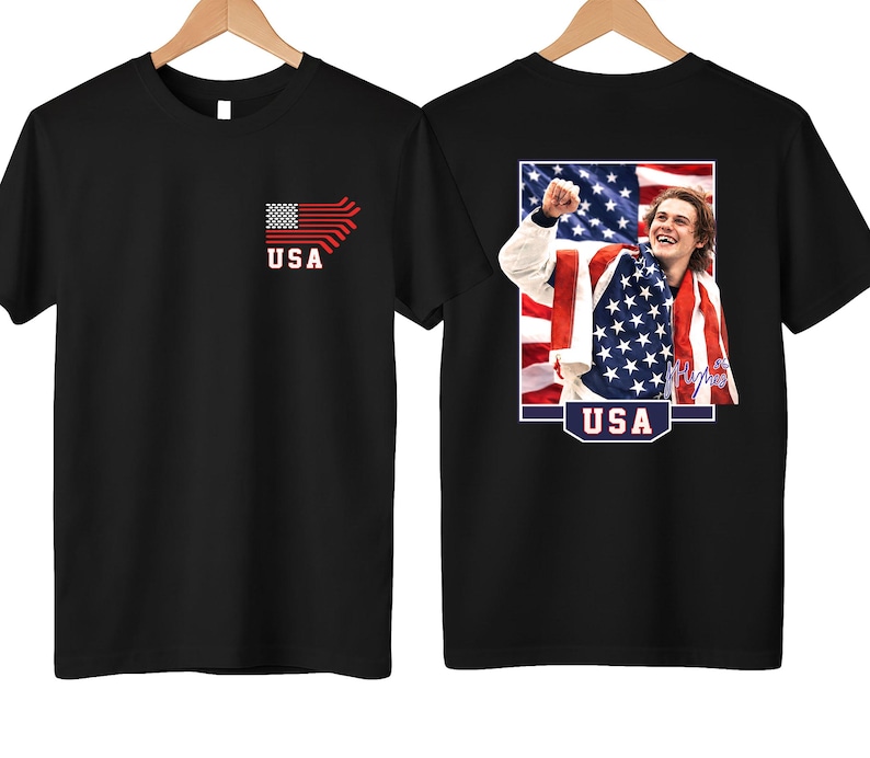 K&ouml;nnte beinhalten: Schwarzes T-Shirt mit einem stilisierten amerikanischen Flaggen-Design und dem Wort "USA" auf der Vorderseite. Auf der R&uuml;ckseite des Shirts befindet sich eine Grafik einer Person, die in eine amerikanische Flagge geh&uuml;llt ist, mit dem Wort "USA" darunter.