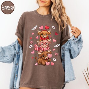 May include: Espresso-colored t-shirt featuring a stacked design of three cartoon Highland cows. The cows are adorned with Valentine's Day themed accessories, including scarves, glasses, and heart-shaped lollipops. Pink hearts and envelopes with hearts are scattered around the cows.