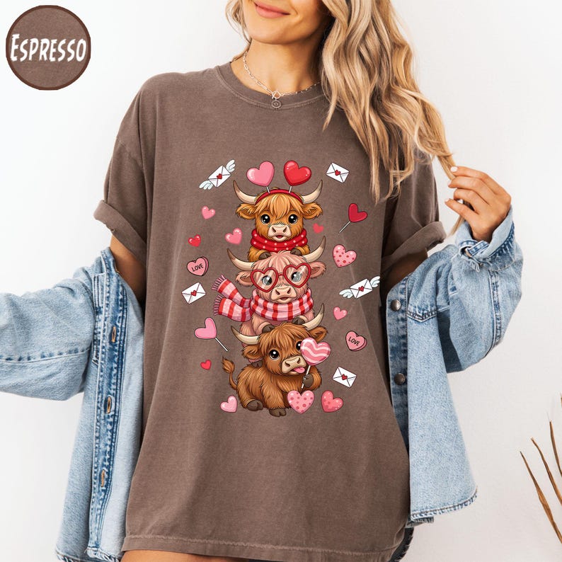 May include: Espresso-colored t-shirt featuring a stacked design of three cartoon Highland cows. The cows are adorned with Valentine's Day themed accessories, including scarves, glasses, and heart-shaped lollipops. Pink hearts and envelopes with hearts are scattered around the cows.