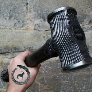 May include: A black metal hammer with a textured head and a wooden handle. The hammer is held in a hand with a tattoo of a snake and a goat.