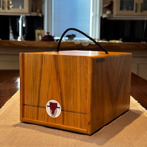 May include: A wooden box with a drawer, featuring a handle and the word "RIHOR" printed on the side. The box has a circular emblem with a red and white design. The box is made of wood with a warm, natural grain.