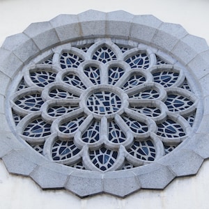 May include: A circular, grey stone rose window with a floral design. The window features petal-shaped openings filled with dark blue glass. The outer frame has a geometric, scalloped edge.
