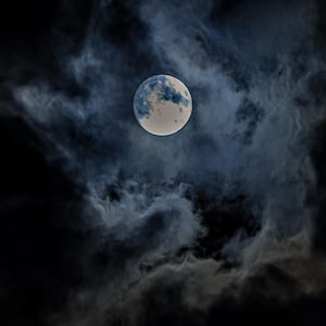 Peut inclure: Une pleine lune est visible dans un ciel nocturne rempli de nuages sombres et tourbillonnants. La lune a une surface claire, blanc cassé, avec des taches bleues et grises. Les nuages sont un mélange de gris foncé et de nuances plus claires.