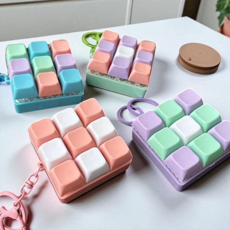 May include: A collection of colorful keyboard keycap keychains in various pastel colors, including pink, blue, green, and purple. Each keychain has a square shape with nine keycaps and a metal ring for attachment.