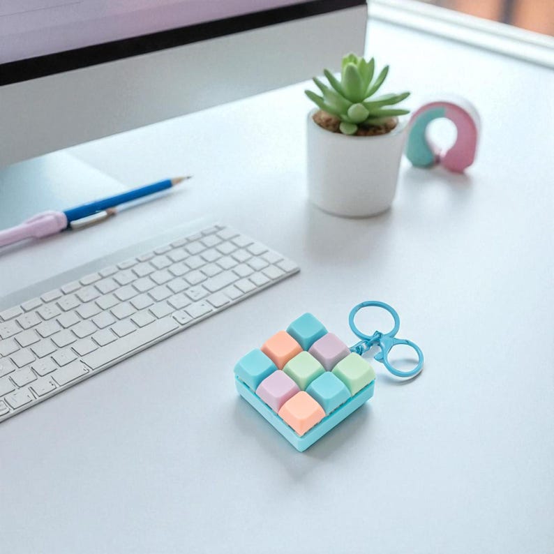 May include: A colorful, square-shaped keychain with a grid of pastel-colored cubes. The keychain has a light blue base and a silver-colored clasp. The cubes are in shades of blue, green, pink, and orange. A keyboard, pencil, and small potted plant are in the background.