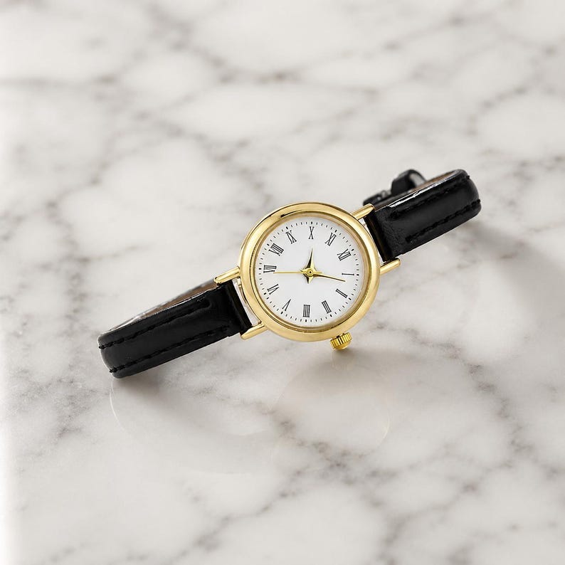 May include: A gold-tone wristwatch with a white face and black leather band. The watch features Roman numeral hour markers and gold hands. The band is approximately 0.5 inches wide. The watch is displayed on a marble surface.