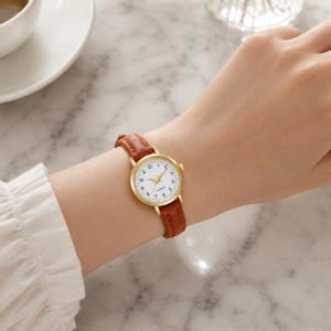 May include: A gold-toned quartz watch with a white face and black numerals. The watch has a brown leather band. The watch is on a wrist.