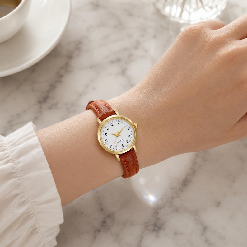 May include: A gold-toned quartz watch with a white face and black numerals. The watch has a brown leather band. The watch is on a wrist.