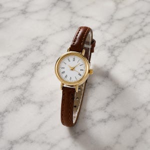 May include: A gold-tone wristwatch with a white face and Roman numeral hour markers. The watch has a brown leather band. The watch face is round and the hands are gold. The watch is a classic style.